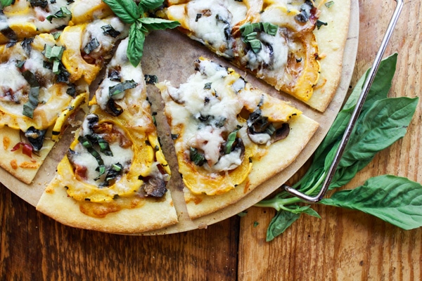 easy thin-crust pizza with roasted veggies Thin-Crust Delicata Squash and Prosciutto Pizza topped with sweet roasted squash, shallots, mushrooms, and melty cheese on top of a pizza stone with fresh basil leaves on the side placed on top of a wooden board.