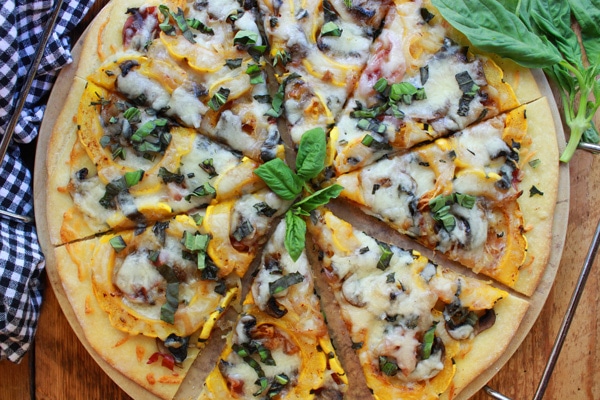 easy thin-crust roasted delicata pizza Thin-Crust Delicata Squash and Prosciutto Pizza topped with sweet roasted squash, shallots, mushrooms, and melty cheese on top of a pizza stone with fresh basil leaves on the side placed on top of a wooden board.
