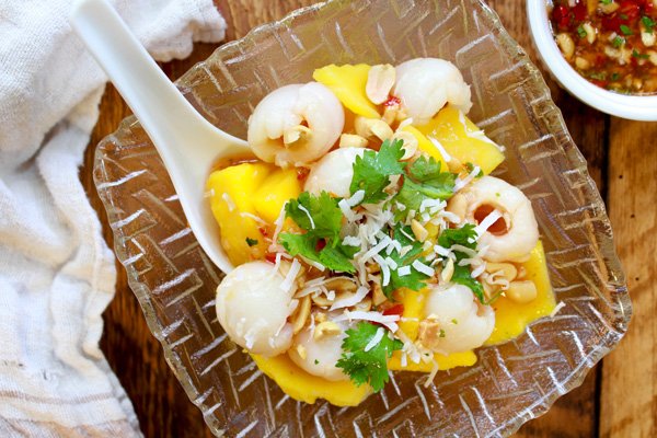 mango salad A tropical mango and lychee salad in a glass serving bowl with a large white spoon inserted on top of a wooden board with a small white bowl of spicy sauce on the side.