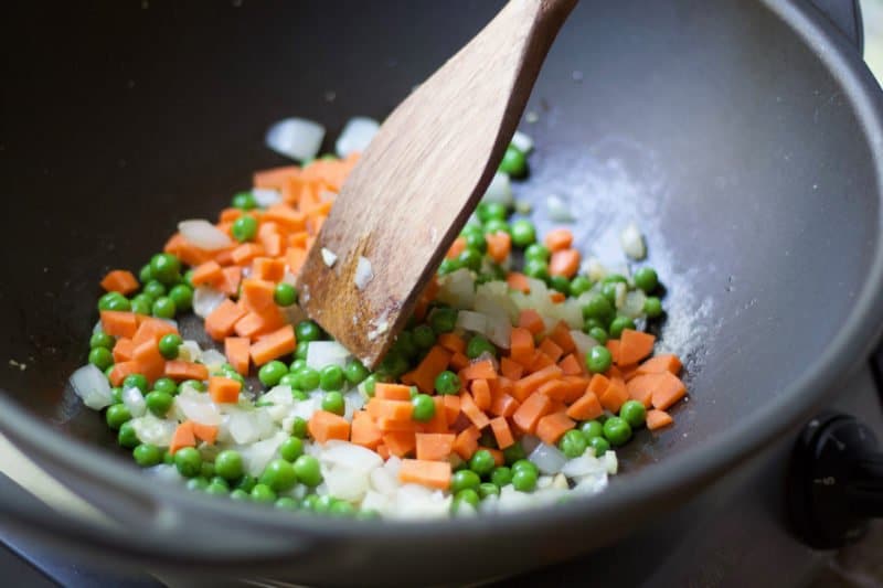 vegetable stir-fry Chopped carrots, onions, and peas being stir-fried in a large wok with a wooden spatula.