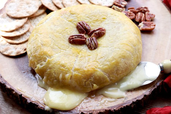 baked brie in puff pastry with oozing cheese A golden-brown baked brie oozing with cheese and topped with pecans right out of the oven placed on top of a wooden board with crackers and pecans placed behind.