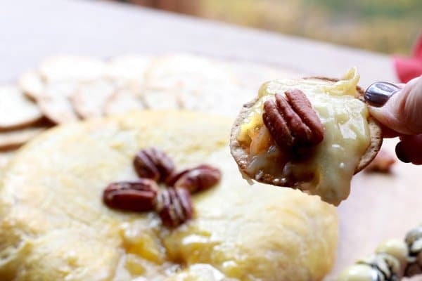 baked brie in puff pastry A person holding a cracker after dipping into a golden-brown baked brie oozing with cheese and topped with pecans.