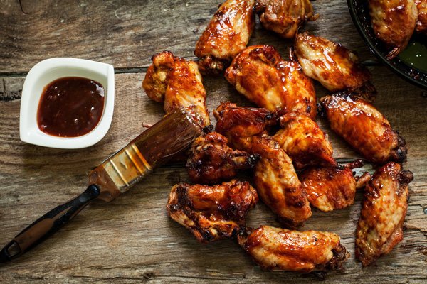 chicken wings Saucy honey garlic chicken wings on top of a wooden board with a side of sauce and a pastry brush.