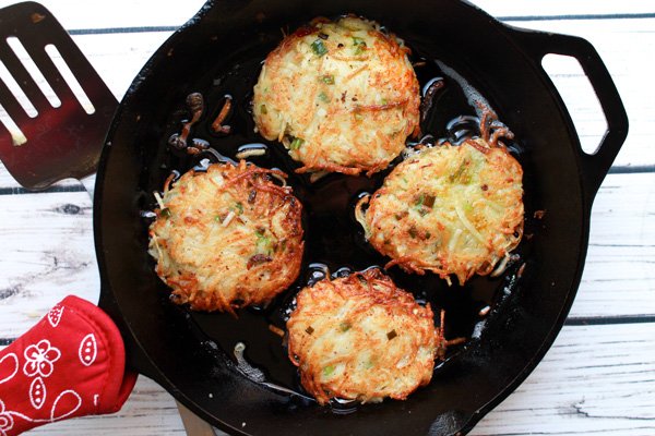 Crispy potato latkes (pancakes) frying in a cast iron skillet with a spatula on the side placed on top of a white wooden board.