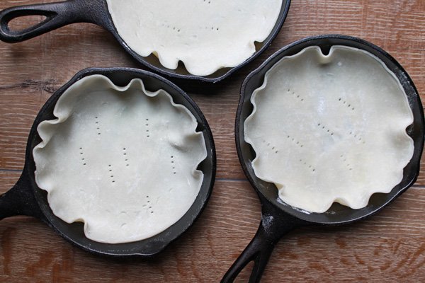 pie crusts Three mini cast iron skillets with raw pastry lined inside on top of a wooden board.