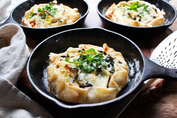 Three golden-brown butternut squash galettes in mini cast iron skillets placed on top of a wooden board with a gray linen napkin and silver pie cutter on the side.