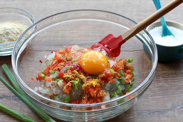 coconut shrimp cakes mixture in bowl A mixture for making coconut shrimp cakes in a large glass mixing bowl with a red rubber spatula inserted on top of a wooden board with a small bowl of salt in the background.