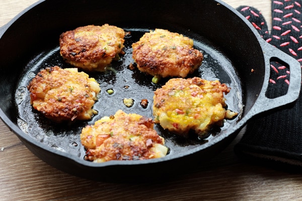 golden brown gluten-free coconut shrimp cakes frying in a skillet Golden-brown coconut shrimp cakes frying up in a cast iron skillet placed on top of a wooden board.