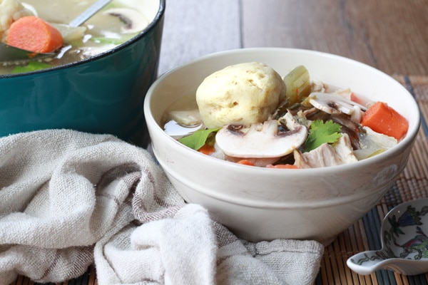 matzo ball soup with Asian flavors A white bowl of matzo ball soup with veggies on top and a soup spoon on the side with a pot of soup in the background placed on top of a bamboo placemat.