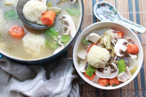 matzo ball soup with Asian flavors A white bowl of matzo ball soup with veggies on top and a soup spoon on the side with a pot of soup in the background placed on top of a bamboo placemat.