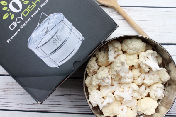 cauliflower florets in Instant Pot steamer basket Cauliflower florets in a silver steamer basket with a box on the side placed on top of a white wooden board.