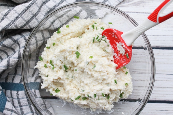 cauliflower mash with cream cheese in a glass bowl Cauliflower mash with chives in a large glass mixing bowl and red spatula inserted placed on top of a white wooden board with a kitchen towel on the side.
