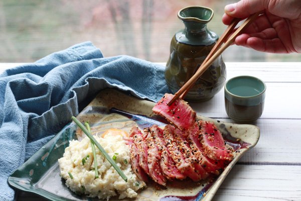 Slices of sesame seared tuna on a square plate with a side of cauliflower mash topped with green onions and a hand picking up a slice of tuna with chopsticks placed on a white board with a sake set and blue napkin in the background.