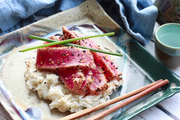 sesame seared tuna on a bed of cheesy cauliflower mash A square plate with slices of sesame seared tuna placed on top of cauliflower mash with a pair of chopsticks on the side.
