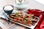 A long white narrow plate with charred Chinese eggplant topped with a garlic pepper sauce with a bowl of white rice and black chopsticks along side placed on top of a bamboo placemat with a red napkin and small teapot on the side.
