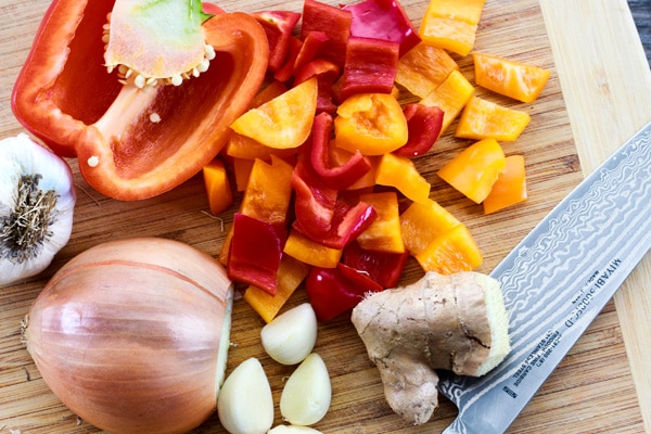 bell peppers, onion, garlic, ginger Colorful red and yellow bell peppers, garlic cloves, onion, and ginger on a wooden cutting board.