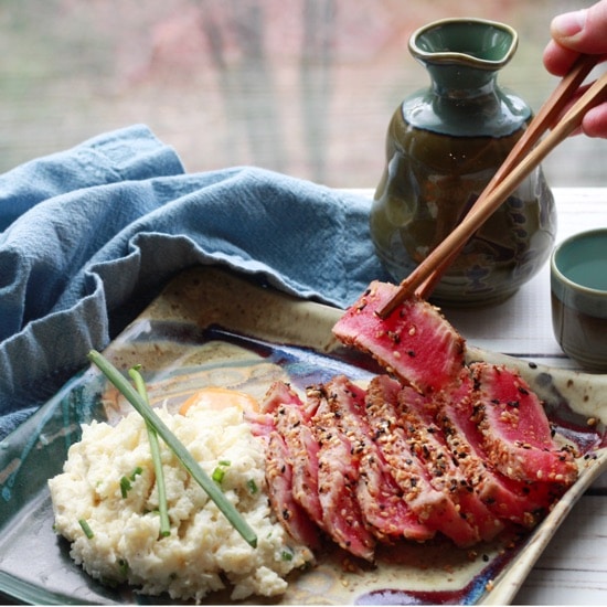 Slices of sesame seared tuna on a square plate with a side of cauliflower mash topped with green onions and a hand picking up a slice of tuna with chopsticks placed on a white board with a sake set and blue napkin in the background.