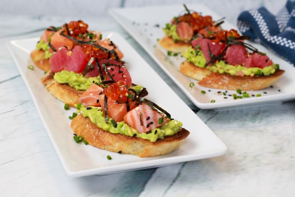 Sushi-grade salmon and tuna are nestled on top of avocado toast presented on a two white plates with a blue napkin on the side.