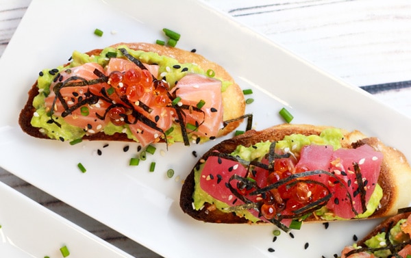 sushi-grade salmon and tuna on avocado toast Sushi-grade salmon and tuna are nestled on top of avocado toast presented on a white plate.