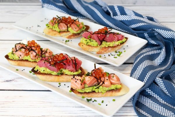Sushi-grade salmon and tuna are nestled on top of avocado toast presented on a two white plates with a blue napkin on the side.