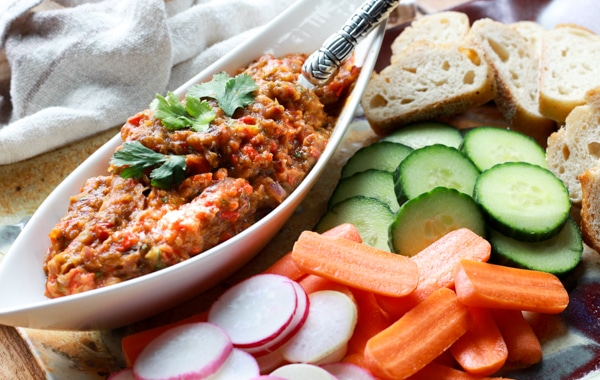 Roasted Chinese eggplant dip in a white boat-shaped bowl with fresh veggies and sliced bread on the side.