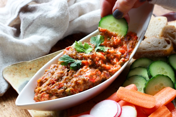 Vibrant, beautiful and delicious Chinese Eggplant Dip is the perfect appetizer, or condiment for any protein! #grilledeggplantdip, #chunkyeggplantdip #roastedeggplant Roasted Chinese eggplant dip in a white boat-shaped bowl with fresh veggies and sliced bread on the side and a hand dipping a cucumber into the dip.
