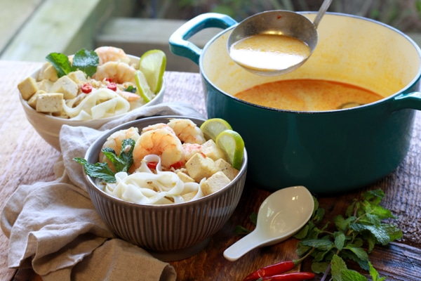 Singapore laksa with shrimp, tofu, and rice noodles in two bowls with a large blue soup pot behind and a white soup spoon on the side, on top of a wooden surface.