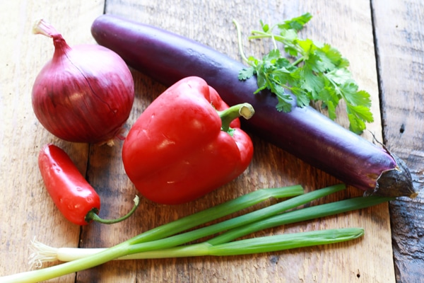 Chinese eggplant, red peppers, red onions, cilantro, and green onions on a wooden board.