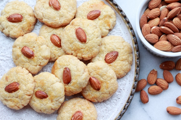 Chinese almond cookies Chinese almond cookies piled on a round white plate with bowl of almonds on the side and extra cookies in front on top of a marble surface.