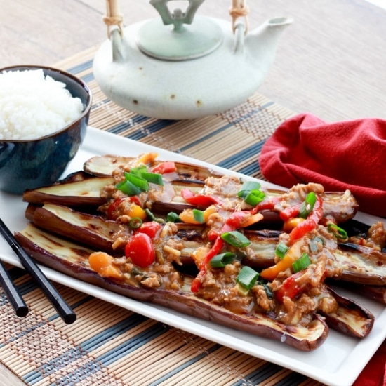 A long white narrow plate with charred Chinese eggplant topped with a garlic pepper sauce with a bowl of white rice and black chopsticks along side placed on top of a bamboo placemat with a red napkin and small teapot on the side.
