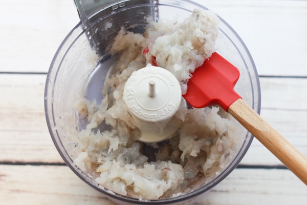 Chopped shrimp in a mini food processor with spatula inserted placed on top of a white wooden board