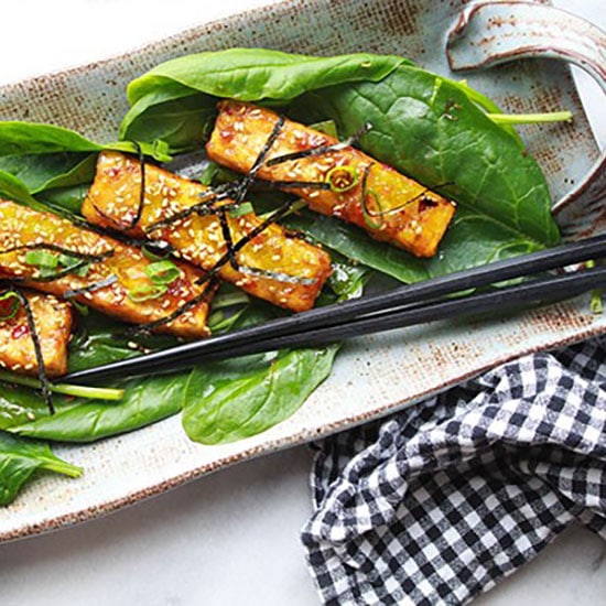 Sesame seared tofu strips on a bed of spinach topped with sesame seeds and seaweed strips presented on a long gray serving plate with black chopsticks on the side.