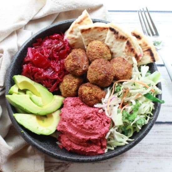 falafel balls in a bowl with beet hummus, slaw, red onions, and avocado on a white board and a fork on the side