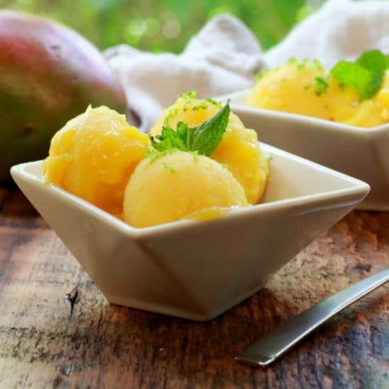 mango coconut sorbet in two white bowls with a spoon and whole mango on the side on top of a wooden board