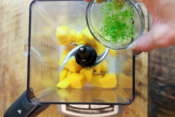 lime zest being placed into a blender with mango chunks
