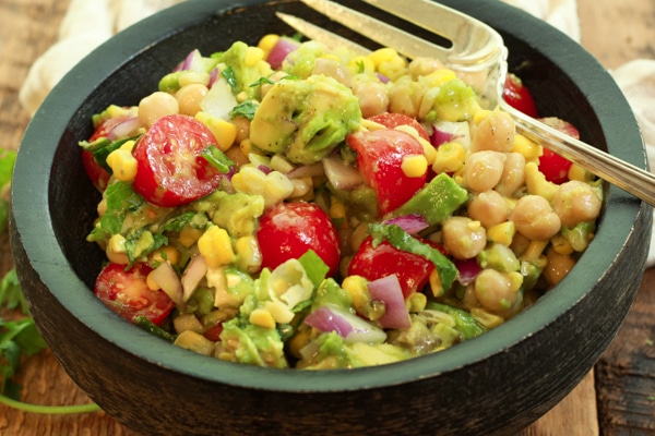 a vibrant corn and avocado salad in a black wooden bowl with a large silver fork and linen napkin the side on top of a wooden board