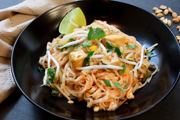 tofu pad thai in a black bowl garnished with a lime wedge with a napkin on the side