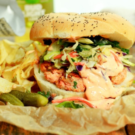 a wild salmon burger topped with broccoli slaw and a spicy aioli on top of parchment paper with a bag of potato chips on the side