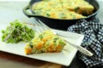 a slice of roasted butternut squash and baby bok choy frittata on a white plate with fresh herbs and a silver fork and checkered napkin on the side and a frittata in a cast iron pan in the background