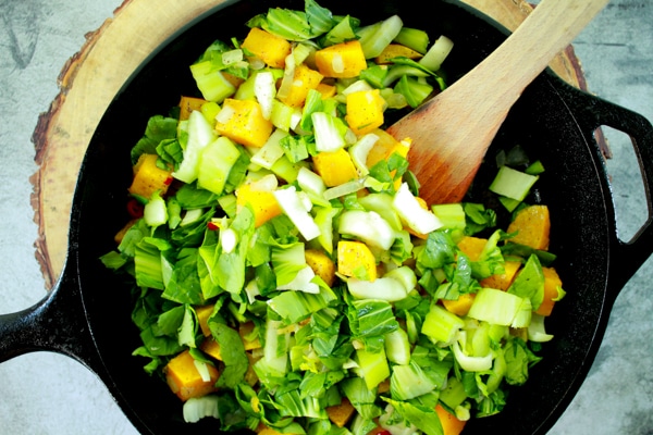 ingredients in a cast iron pan for a roasted butternut squash and bok choy frittata with a wooden spatula inserted in the pan