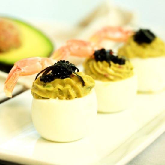 avocado deviled eggs topped with fresh shrimp and caviar stacked on a white plate with a halved avocado in the background.