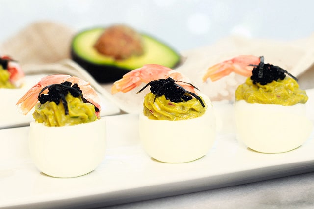 avocado deviled eggs topped with fresh shrimp and caviar stacked on a white plate with a halved avocado in the background.