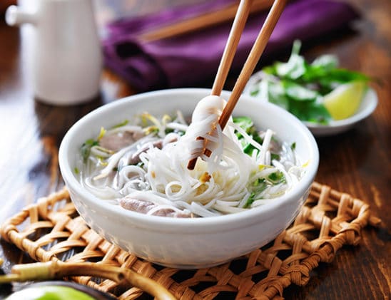 wooden chopsticks picking up noodles out of a white soup bowl of Vietnamese pho ga soup on top of a wicker placemat with garnishings on the side
