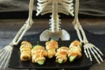 Scary Halloween Jalapeño Popper Mummies on a black plate with a skeleton sitting behind.