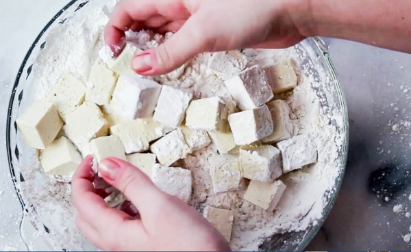 a woman tossing tofu cubes into corn starch in a glass dish