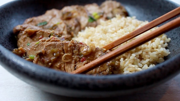 slices of pork tenderloin in a black bowl with a side of brown rice and wooden chopsticks