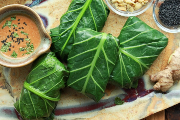 tofu collard wraps on a platter with peanut sauce being poured on top and extra sauce in a small bowl on the side along with a bowl of chopped peanuts and a bulb of ginger