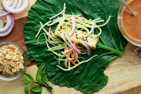 a large collard green leaf with crispy tofu and broccoli slaw on top and peanut sauce in a bowl on the side along with a bowl of chopped peanuts