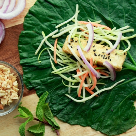 a large collard green leaf with crispy tofu and broccoli slaw on top and peanut sauce in a bowl on the side along with a bowl of chopped peanuts