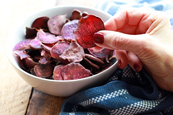 woman holding a purple sweet potato chip with a white bowl of chips on a board and a blue napkin along side